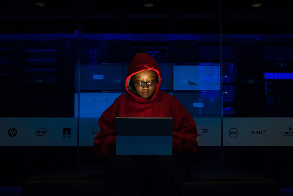 Person in red hoodie working on a laptop in a blue-lit server room,illustrating increased risk of vitamin D deficiency from indoor life.
