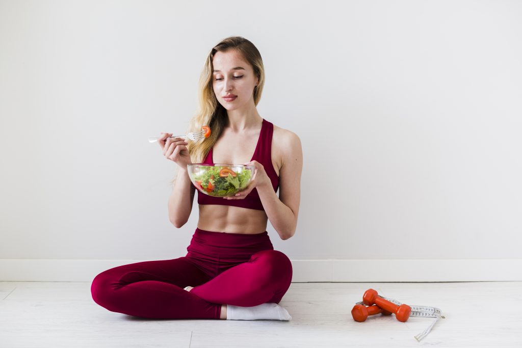 Fit woman eating fresh salad next to a dumbbell, promoting a balanced diet for weight loss with PCOS.
