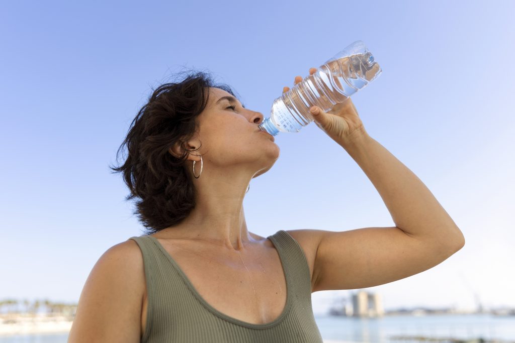 A woman drinking water outdoors on a hot day, emphasizing the role of hydration in curbing overeating.