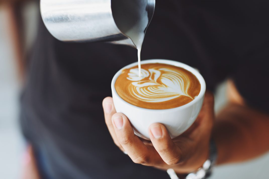 A person pours milk into a steaming cup of coffee, which can trigger a migraine.
