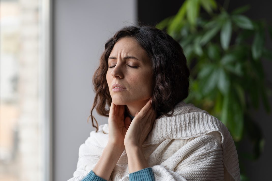 Woman in a white shawl holding her throat, eyes closed in discomfort, feeling a sore throat relief from raw honey.