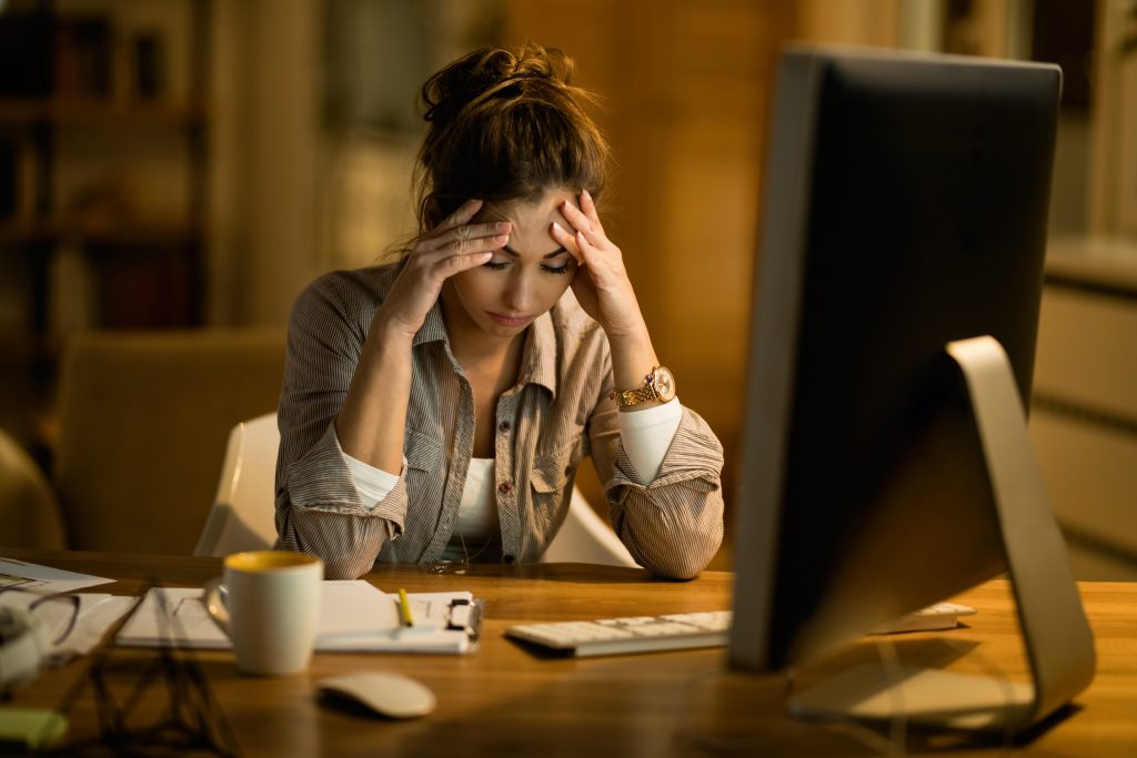 A woman holding her head at night, surrounded by a laptop, tea cup, and writing pad, reflecting stress management.